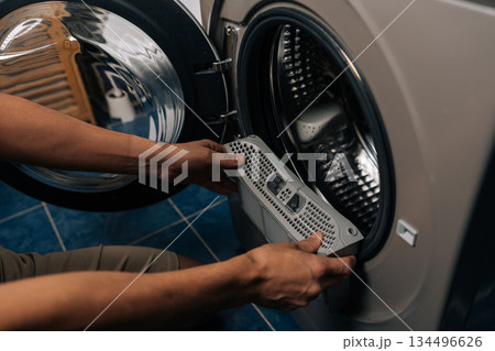 Close-up of person inspecting and removing lint filter from front-loading washing machine or tumble dryer, ensuring proper cleaning and essential appliance maintenance for optimal performance. Close-up of person inspecting and removing lint filter from front-loading washing machine or tumble dryer, ensuring proper cleaning and essential appliance maintenance for optimal performance. 134496626