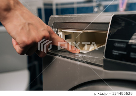 Hand reaching for detergent dispenser on washing machine, indicating process of preparing laundry cycle with cleaning products in household setting. Concept of household chores. Hand reaching for detergent dispenser on washing machine, indicating process of preparing laundry cycle with cleaning products in household setting. Concept of household chores. 134496629