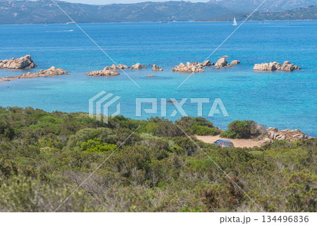 A bright coastal landscape with turquoise water, rocky formations and dense shrubs along the shore. The peaceful Mediterranean atmosphere and clear summer sky create a warm, inviting seaside scenery. 134496836