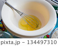broken raw eggs in a white bowl with a whisk for whipping on a multi-colored tablecloth 134497310