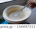 the finished pancake batter in a white bowl with a metal whisk holds the woman's hand of an elderly woman 134497313