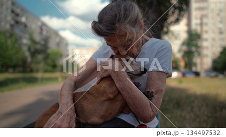 Senior 90-year-old woman with gray hair and deep wrinkles sits outdoors In assisted living facility on bench with small dachshund dog. Old female hugs and cuddles pet in the park on a bench 134497532