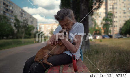Senior 90-year-old woman with gray hair and deep wrinkles sits outdoors In assisted living facility on bench with small dachshund dog. Old female hugs and cuddles pet in the park on a bench 134497582