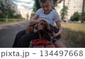 Senior 90-year-old woman with gray hair and deep wrinkles sits outdoors In assisted living facility on bench with small dachshund dog. Old female hugs and cuddles pet in the park on a bench 134497668