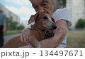 Senior 90-year-old woman with gray hair and deep wrinkles sits outdoors In assisted living facility on bench with small dachshund dog. Old female hugs and cuddles pet in the park on a bench 134497671