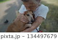 Senior 90-year-old woman with gray hair and deep wrinkles sits outdoors In assisted living facility on bench with small dachshund dog. Old female hugs and cuddles pet in the park on a bench 134497679