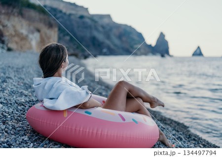 Woman, beach, ocean. Young woman relaxes on pebble beach with donut float, gazing at calm ocean, perfect for tranquil summer travel. 134497794