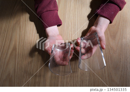 A top view of a child's hands holding two broken pieces of a clear glass on a wooden surface. Conceptual concepts: safety, household accidents, child safety, parental control. A top view of a child's hands holding two broken pieces of a clear glass on a wooden surface. Conceptual concepts: safety, household accidents, child safety, parental control. 134501335