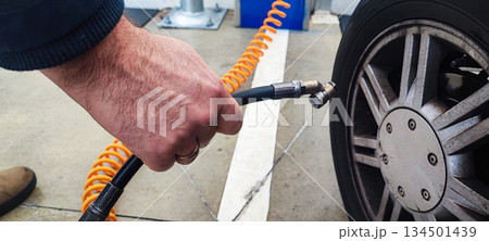 A close-up of an adult's hand inflating a car tire with a compressed air hose at a gas station or garage. Concept: vehicle maintenance, car care, road safety. A close-up of an adult's hand inflating a car tire with a compressed air hose at a gas station or garage. Concept: vehicle maintenance, car care, road safety. 134501439