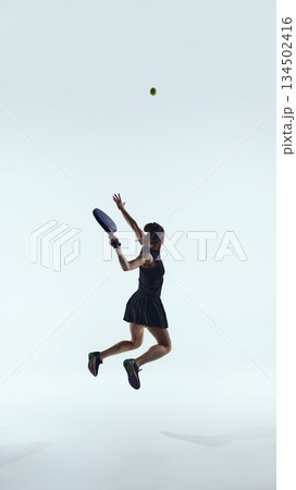 Female padel player jumps mid-air to hit the ball against clean studio background. Female padel player jumps mid-air to hit the ball against clean studio background. 134502416