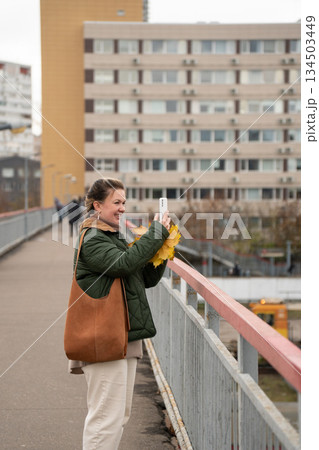 Woman in a green jacket stands on a pedestrian bridge, holding a smartphone and yellow autumn leaves against an urban residential background 134503449