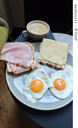 Simple breakfast plate with fried eggs, ham and cheese sandwiches, and a cup of coffee on a small table. The image represents morning routine, home comfort, and everyday food lifestyle concept 134504049