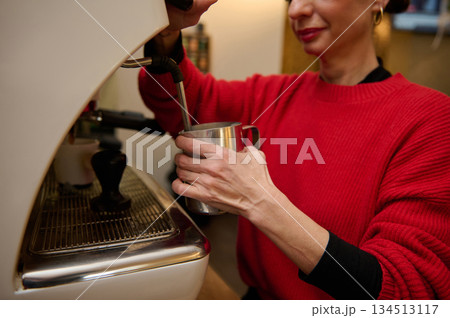 A cafe owner in a red sweater pours milk from a metal pitcher into a steaming espresso machine, conveying hospitality, skill, and daily cafe operation. A cafe owner in a red sweater pours milk from a metal pitcher into a steaming espresso machine, conveying hospitality, skill, and daily cafe operation. 134513117