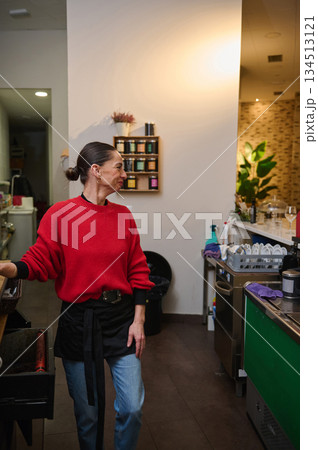A smiling cafe owner in a red knit sweater and apron greets guests near the counter in a warm, busy cafe with shelves, plants, and dishes. A smiling cafe owner in a red knit sweater and apron greets guests near the counter in a warm, busy cafe with shelves, plants, and dishes. 134513121