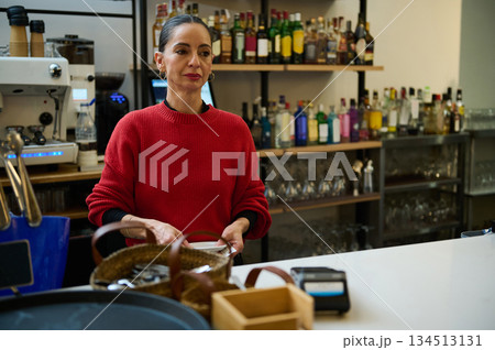 Hispanic cafe owner in a red sweater stands behind a busy bar, preparing orders. Shelves of bottles and glassware create a warm, welcoming hospitality scene. 134513131