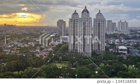 A glowing golf course with sand bunkers and floodlights stretches beneath ornate towers and a vivid sunset, blending sport, architecture, and nature in a dramatic dusk cityscape in Jakarta A glowing golf course with sand bunkers and floodlights stretches beneath ornate towers and a vivid sunset, blending sport, architecture, and nature in a dramatic dusk cityscape in Jakarta 134518858