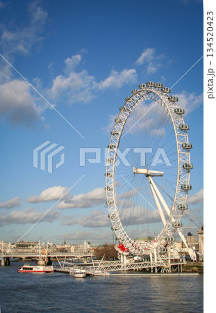 view of the London Eye on the River Thames under a clear blue sky, with clouds and a city skyline along the riverbank, suggesting travel, tourism and urban imagery view of the London Eye on the River Thames under a clear blue sky, with clouds and a city skyline along the riverbank, suggesting travel, tourism and urban imagery 134520423