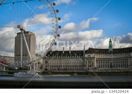 view of the London Eye on the River Thames under a clear blue sky, with clouds and a city skyline along the riverbank, suggesting travel, tourism and urban imagery 134520424