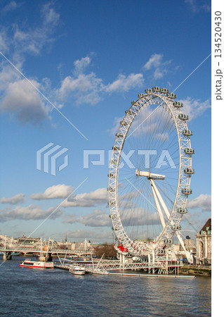 view of the London Eye on the River Thames under a clear blue sky, with clouds and a city skyline along the riverbank, suggesting travel, tourism and urban imagery view of the London Eye on the River Thames under a clear blue sky, with clouds and a city skyline along the riverbank, suggesting travel, tourism and urban imagery 134520430