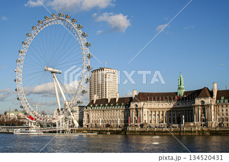 view of the London Eye on the River Thames under a clear blue sky, with clouds and a city skyline along the riverbank, suggesting travel, tourism and urban imagery 134520431