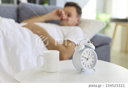 Sleepy man lying near alarm clock in bed, morning wake-up with coffee mug Sleepy man lying near alarm clock in bed, morning wake-up with coffee mug 134523321