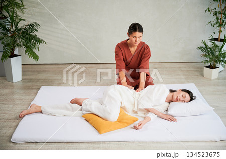 Masseuse making thai yoga massage. Woman therapist doing traditional massage treatment for female patient indoors. Client lying on mat, meditating, enjoying hand massage in calm atmosphere. Masseuse making thai yoga massage. Woman therapist doing traditional massage treatment for female patient indoors. Client lying on mat, meditating, enjoying hand massage in calm atmosphere. 134523675