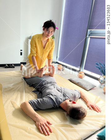 Masseuse making thai yoga massage. Woman therapist doing traditional massage treatment, stretching male patient's body. Client lying on mat, enjoying hand massage in calm atmosphere. 134523683