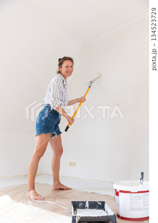 Portrait young adult beautiful single happy woman painting wall paint roller brush white color living room moving new home flat . Female painter decorating interior renovation residential apartment 134523729