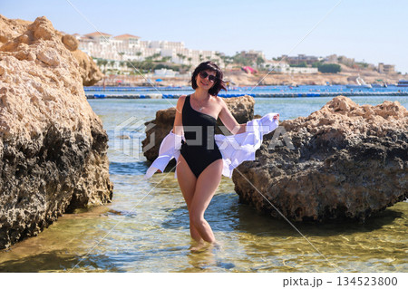 A woman poses on the beach standing in the water near a large rock 134523800