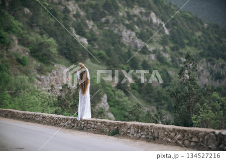 A woman stands on a stone wall overlooking a mountain. She is wearing a white dress and she is in a contemplative mood. A woman stands on a stone wall overlooking a mountain. She is wearing a white dress and she is in a contemplative mood. 134527216