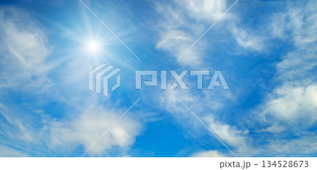 Bright sun shining in a clear blue sky with white clouds. 134528673