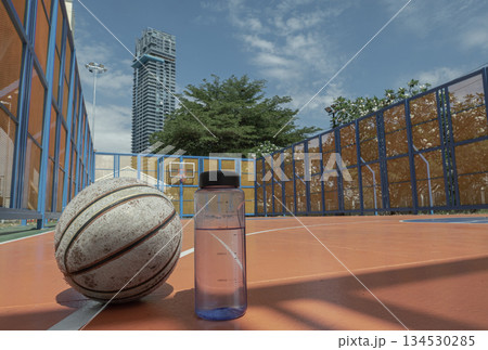 The bright blue sky and soaring modern building stand in stark relief to the grounded, worn basketball and the unassuming water bottle, all resting quietly on the vibrant orange court. The bright blue sky and soaring modern building stand in stark relief to the grounded, worn basketball and the unassuming water bottle, all resting quietly on the vibrant orange court. 134530285