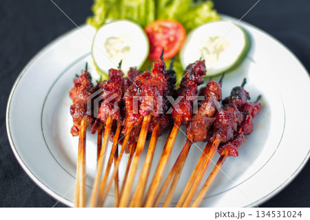 Sate Rembiga, traditional spicy grilled beef skewers from Lombok, Indonesia, served on a white plate with fresh vegetable garnish. 134531024