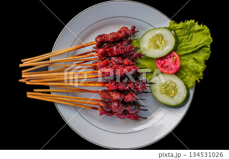Sate Rembiga, traditional spicy skewered beef or mutton meat from Lombok, Indonesia, served on white plate with fresh cucumber and tomato garnish on black background. 134531026