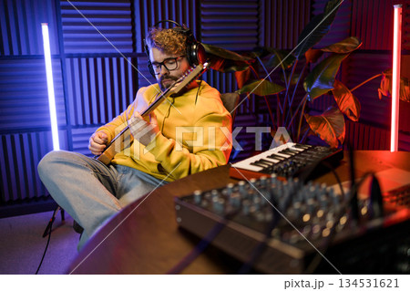 A focused male musician fine-tunes a guitar against the backdrop of a digital sound editing setup in a contemporary recording studio environment. A focused male musician fine-tunes a guitar against the backdrop of a digital sound editing setup in a contemporary recording studio environment. 134531621