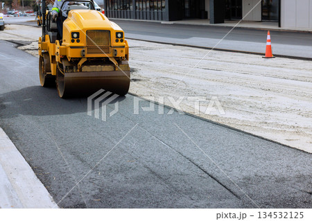 Steamroller roller compacts freshly laid asphalt on street under construction in bustling city setting. 134532125