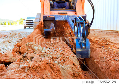 Worker operates trenching machine to create trench in dirt at construction site. Worker operates trenching machine to create trench in dirt at construction site. 134532129