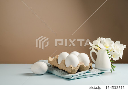 Easter eggs and flowers. White eggs in cardboard box and Freesia white spring bouquet in a ceramic jug Easter eggs and flowers. White eggs in cardboard box and Freesia white spring bouquet in a ceramic jug 134532813