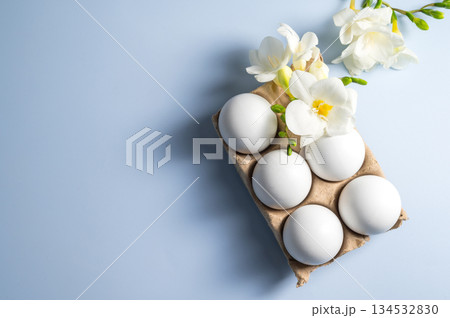 Easter eggs and flowers. White eggs in cardboard box and Freesia white spring flower, flat lay on blue background with copy space Easter eggs and flowers. White eggs in cardboard box and Freesia white spring flower, flat lay on blue background with copy space 134532830
