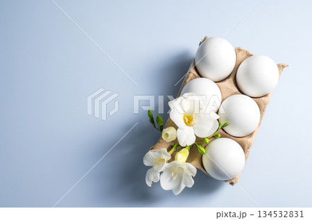 Easter eggs and flowers. White eggs in cardboard box and Freesia white spring flower, flat lay on blue background with copy space 134532831