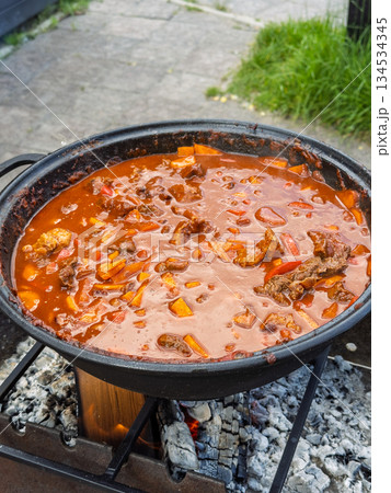 Communal outdoor feast featuring robust stew of meat and peppers slow cooked in heavy pot over charcoal flame 134534345