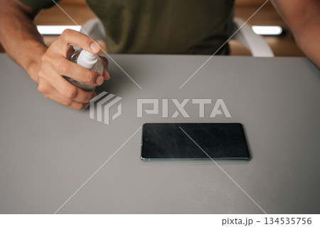 Close-up of male hands spraying sanitizer onto mobile phone, highlighting importance of hygiene and disinfection in everyday life on gray table surface. Concept of mobile phone maintenance and service 134535756