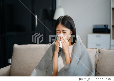 Asian woman suffering from flu symptoms, covered with a blanket on a sofa. Concept of illness, healthcare, and recovery. 134539650