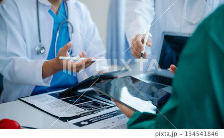 Medical team having a meeting with doctors in white lab coats and surgical scrubs seated at a table discussing a patients working online using computers 134541846
