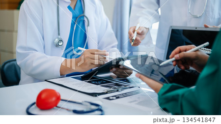 Medical team having a meeting with doctors in white lab coats and surgical scrubs seated at a table discussing a patients working online using computers 134541847