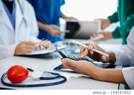 Medical team having a meeting with doctors in white lab coats and surgical scrubs seated at a table discussing a patients working online using computers 134541852