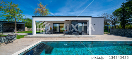Modern modular house backyard featuring a rectangular swimming pool, large patio with wooden sun loungers, and umbrellas against a blue sky. Contemporary prefabricated home design 134543426