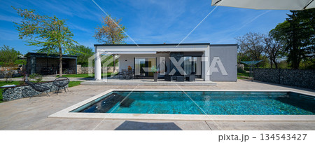 Modern modular house backyard featuring a rectangular swimming pool, large patio with wooden sun loungers, and umbrellas against a blue sky. Contemporary prefabricated home design Modern modular house backyard featuring a rectangular swimming pool, large patio with wooden sun loungers, and umbrellas against a blue sky. Contemporary prefabricated home design 134543427