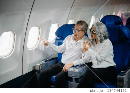 Senior Asian couple capturing memories on a plane, blending travel with technology. Smiling, stylish, Senior Asian couple capturing memories on a plane, blending travel with technology. Smiling, stylish, 134544121