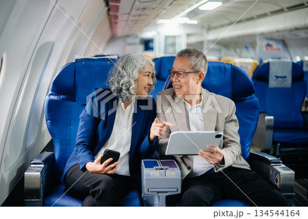 Senior Asian couple capturing memories on a plane, blending travel with technology. Smiling, stylish, Senior Asian couple capturing memories on a plane, blending travel with technology. Smiling, stylish, 134544164